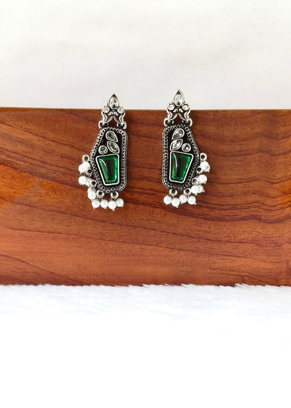 A pair of silver-toned traditional Indian jhumka earrings with green stone inlays and dangling pearl-like beads, displayed on a wooden block against a white background.