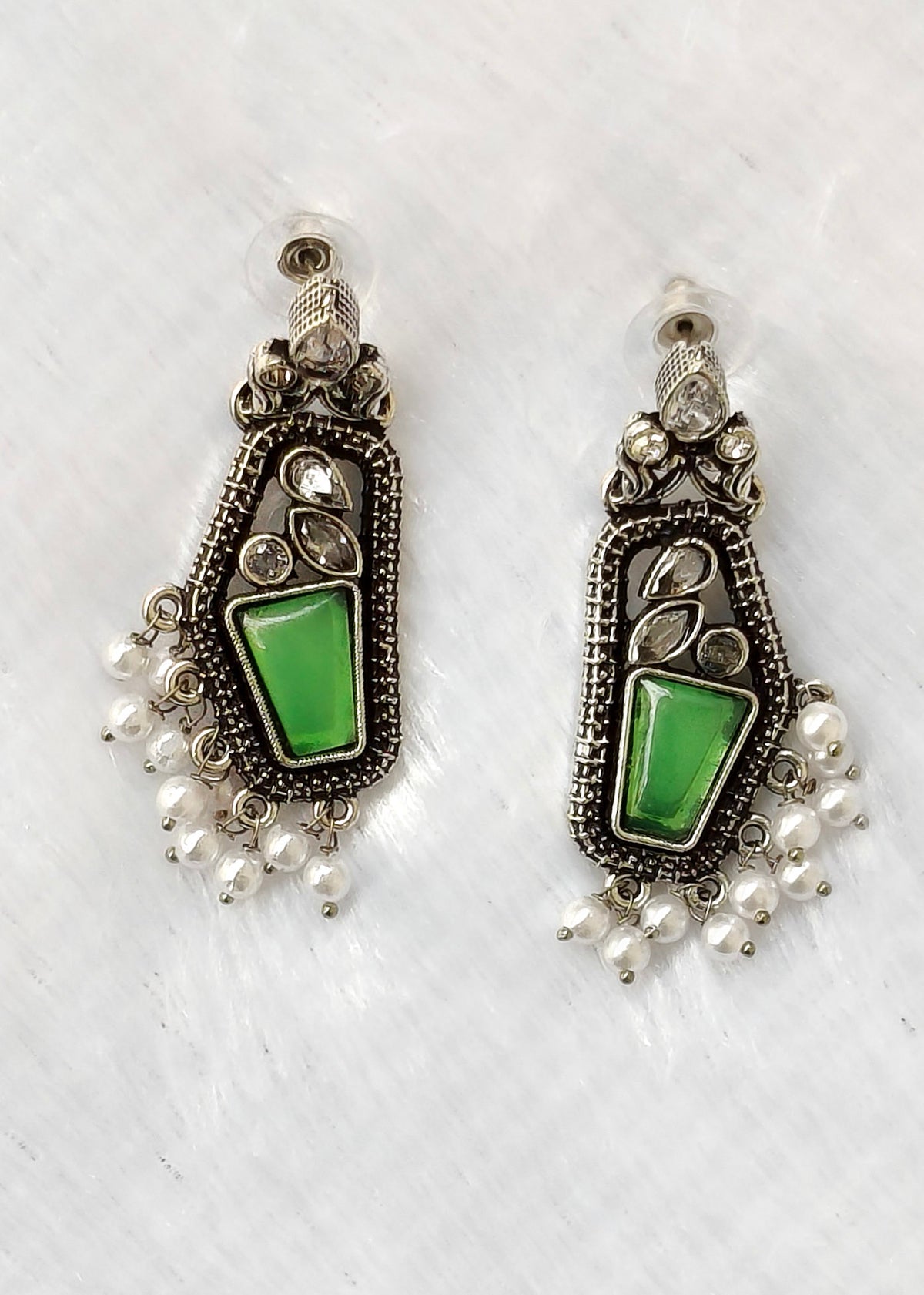 A pair of silver-toned dangle earrings with green stone inlays and dangling pearl-like beads, displayed on a white textured fabric background.