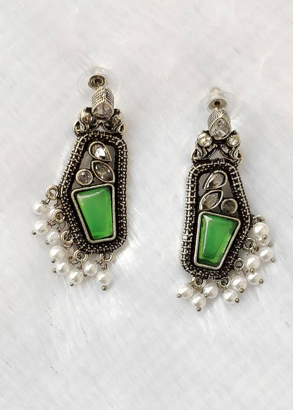 A pair of silver-toned dangle earrings with green stone inlays and dangling pearl-like beads, displayed on a white textured fabric background.