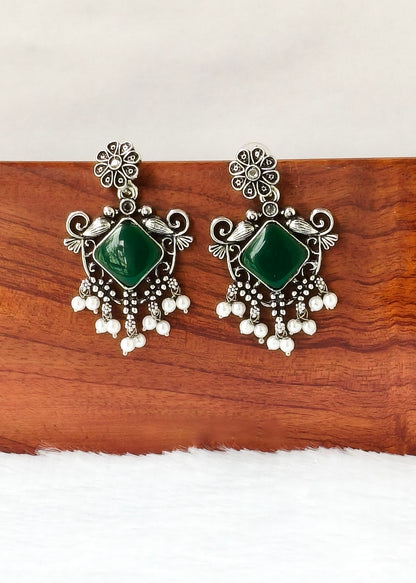 A pair of ornate silver-toned earrings with green square stones and dangling pearl-like beads, displayed on a wooden block against a white background.