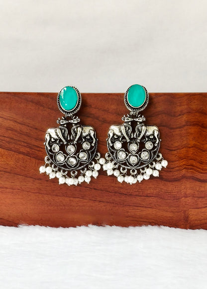 A pair of ornate silver-toned jhumka earrings with oval turquoise green stones and dangling white bead chains, displayed on a wooden block against a white background.