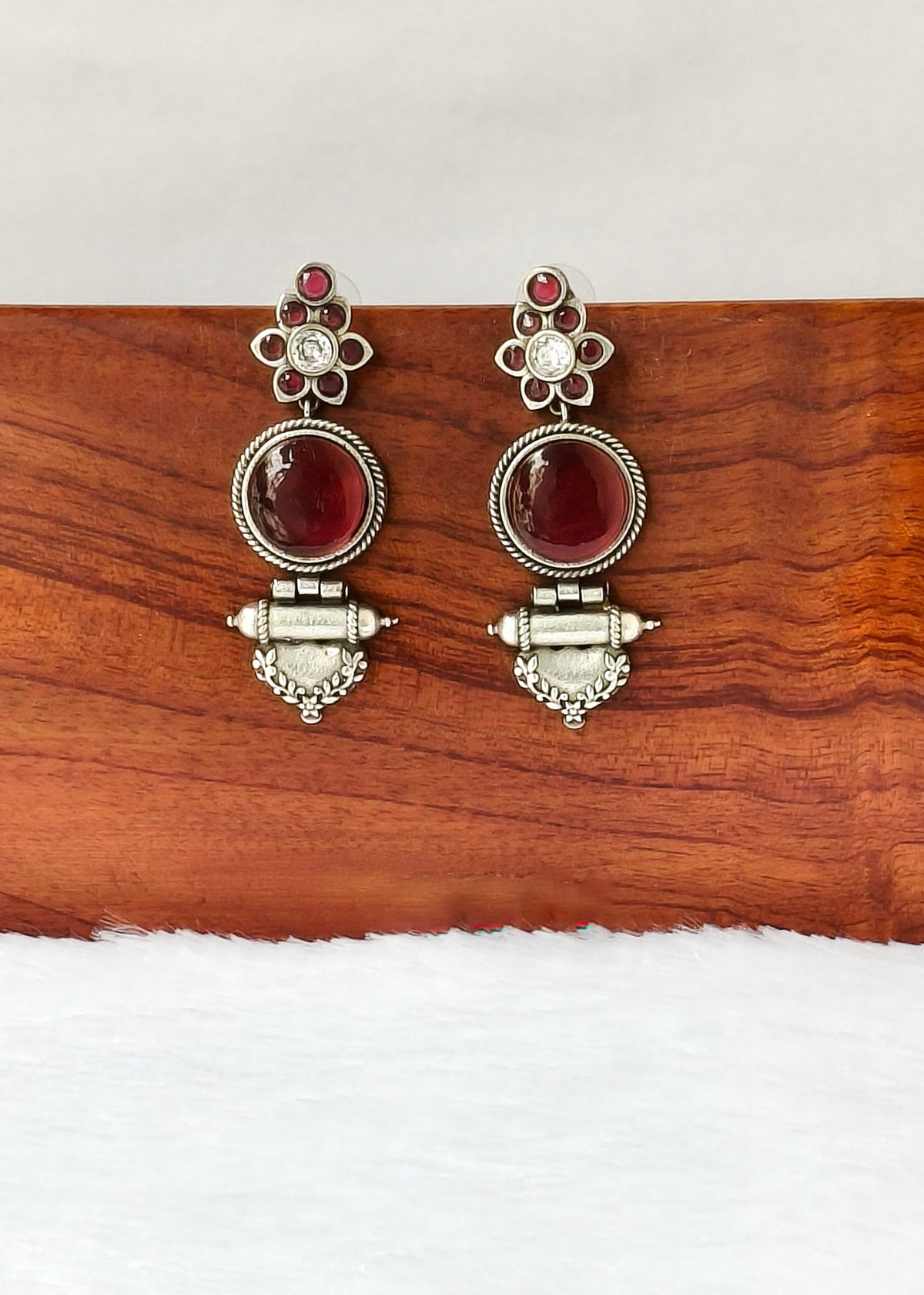 A pair of silver-toned traditional dangle earrings with oval red gemstones and small clear stones, displayed on a wooden block against a white background.