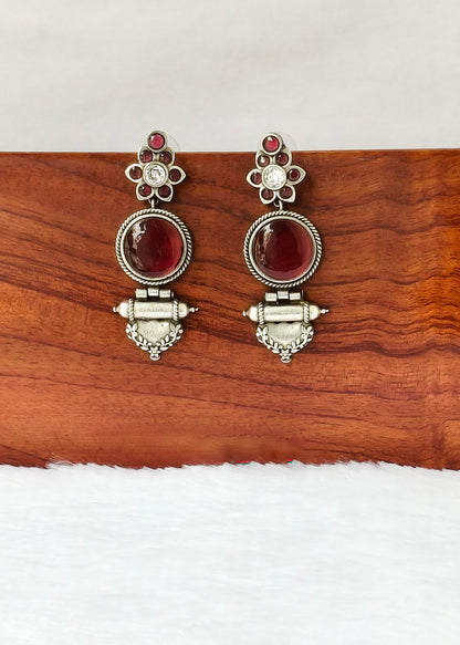 A pair of silver-toned traditional dangle earrings with oval red gemstones and small clear stones, displayed on a wooden block against a white background.