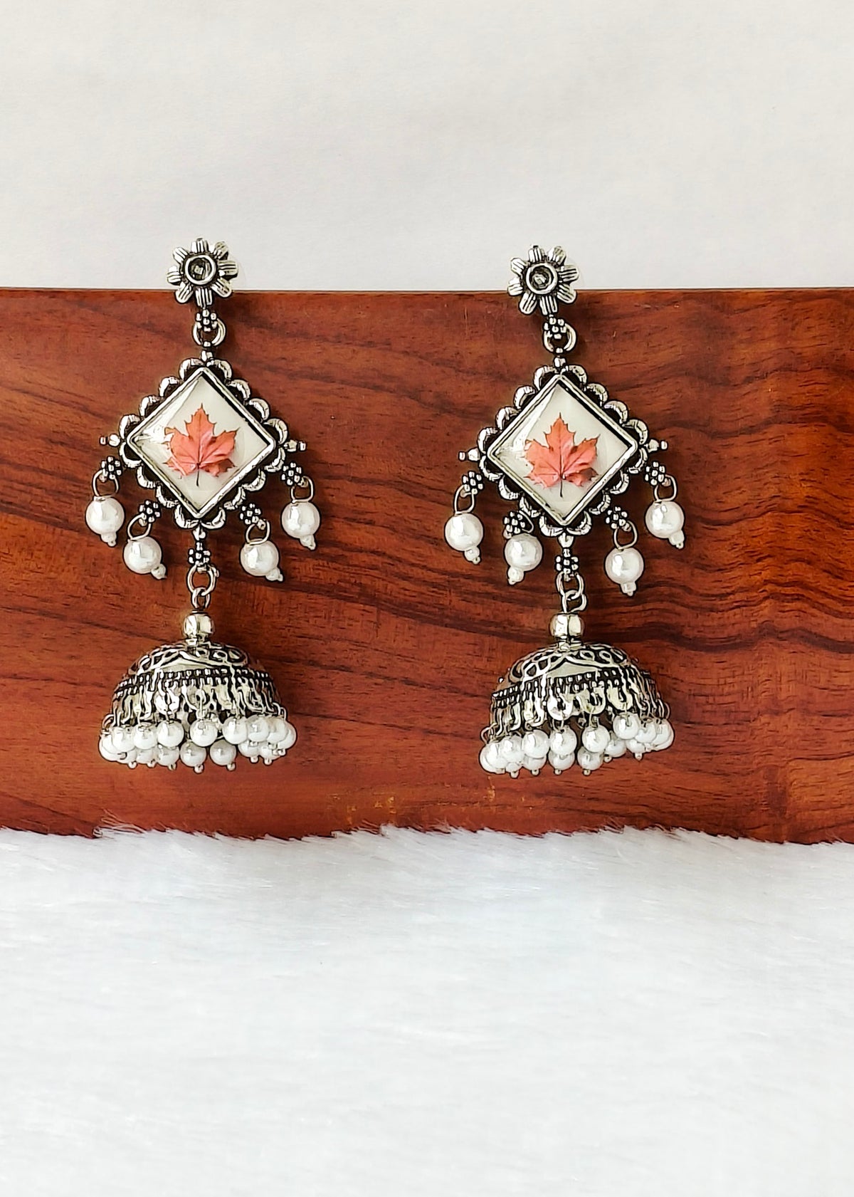 A pair of traditional Indian silver-toned jhumka earrings with white pearl bead drops and a central square inlay featuring a coral-pink maple leaf on a white background, displayed on a wooden block.