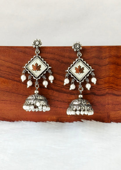 A pair of silver-toned Indian jhumka earrings with white pearl bead drops and a central white square inlay featuring a small brown maple leaf, displayed on a wooden block against a white background.
