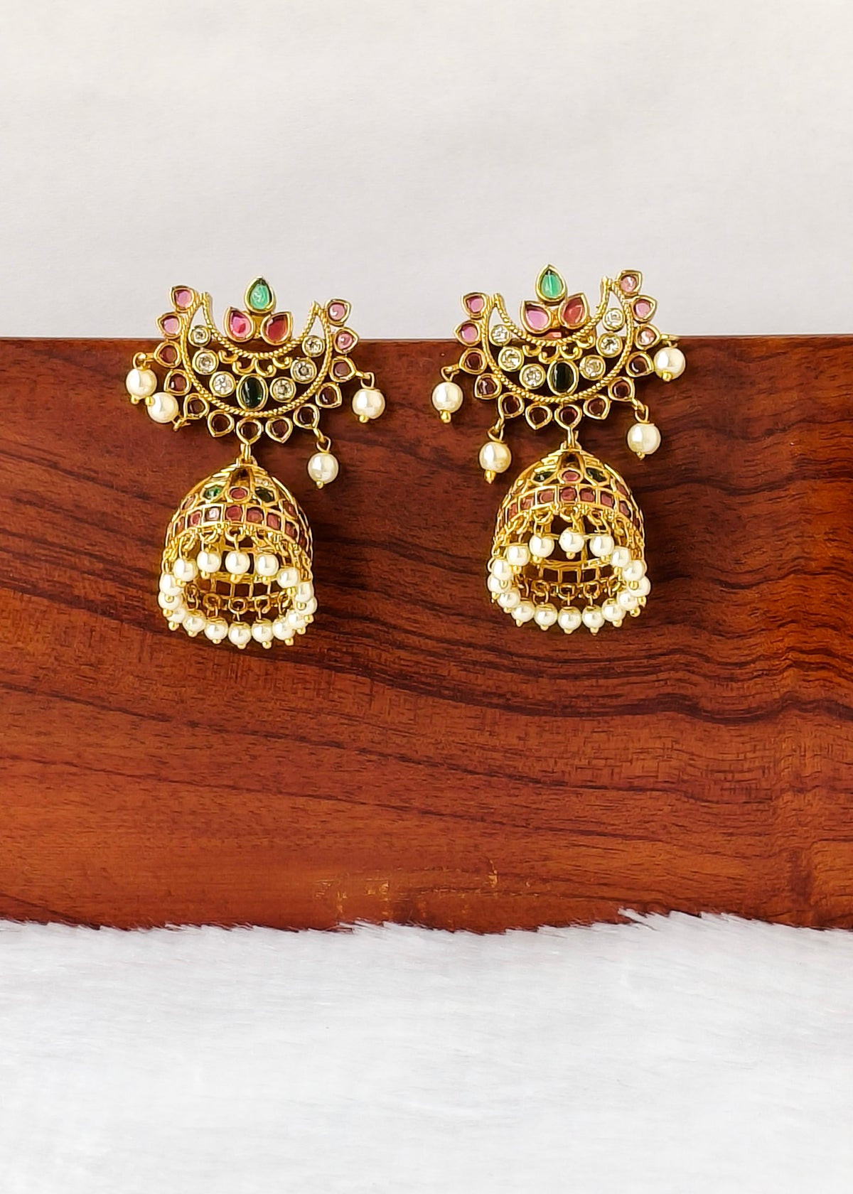 A pair of ornate gold-toned Indian jhumka earrings with red, green, and white stone accents and dangling pearl-like beads, displayed on a wooden block against a white background.