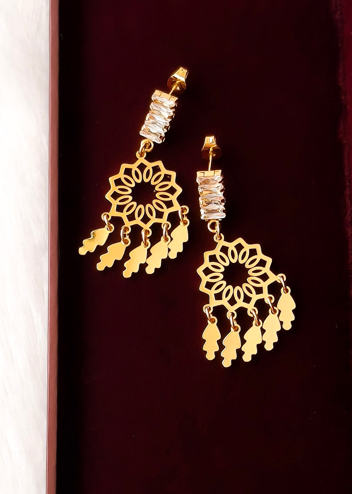 A pair of gold-toned dangle earrings with intricate filigree cutouts, clear rhinestone-studded posts, and dangling leaf-shaped charms, displayed on a dark burgundy background.