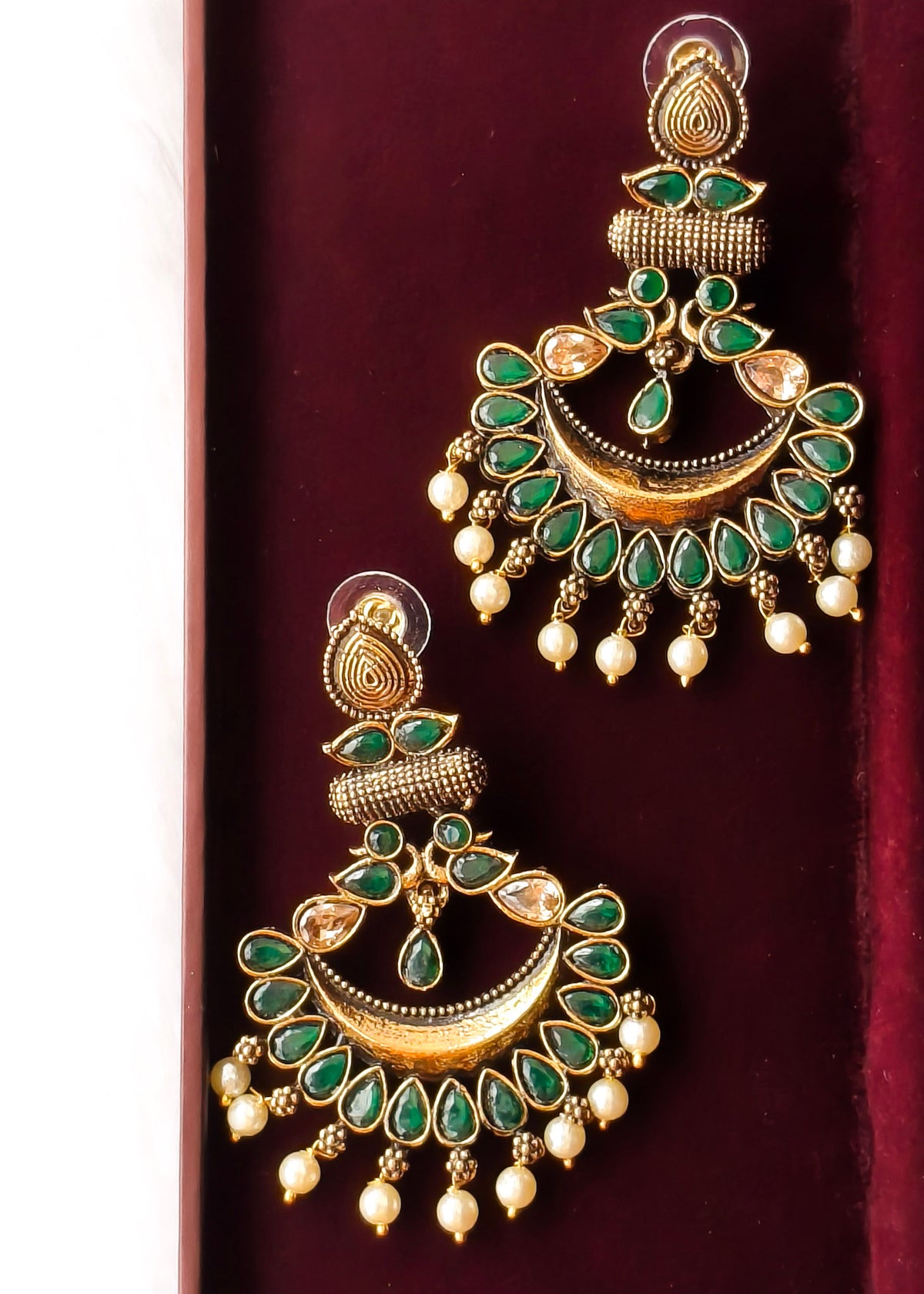 A pair of gold-toned Indian jhumka earrings embellished with green stones, clear rhinestones, and white pearl-like drops, displayed on a dark red velvet surface.