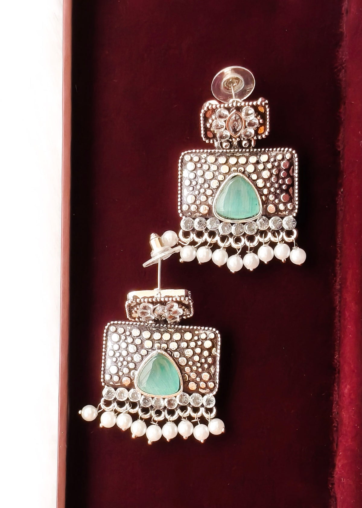 A pair of silver-toned Indian jhumka earrings with green teardrop stones and white bead fringe, displayed on a dark red velvet surface.