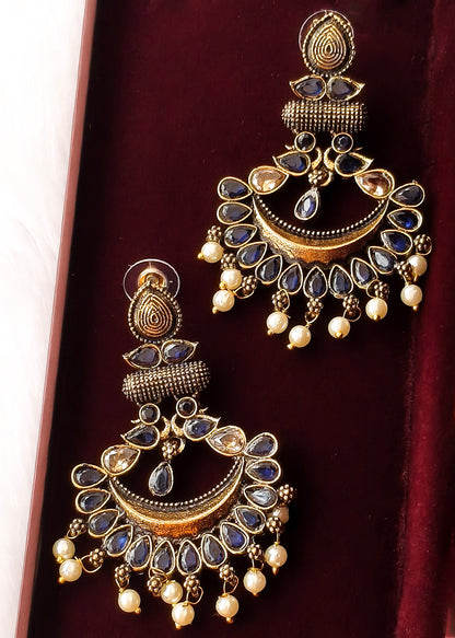 A pair of ornate gold-toned Indian chandelier earrings with blue stone accents and pearl-like dangling beads, displayed on a dark red velvet surface.