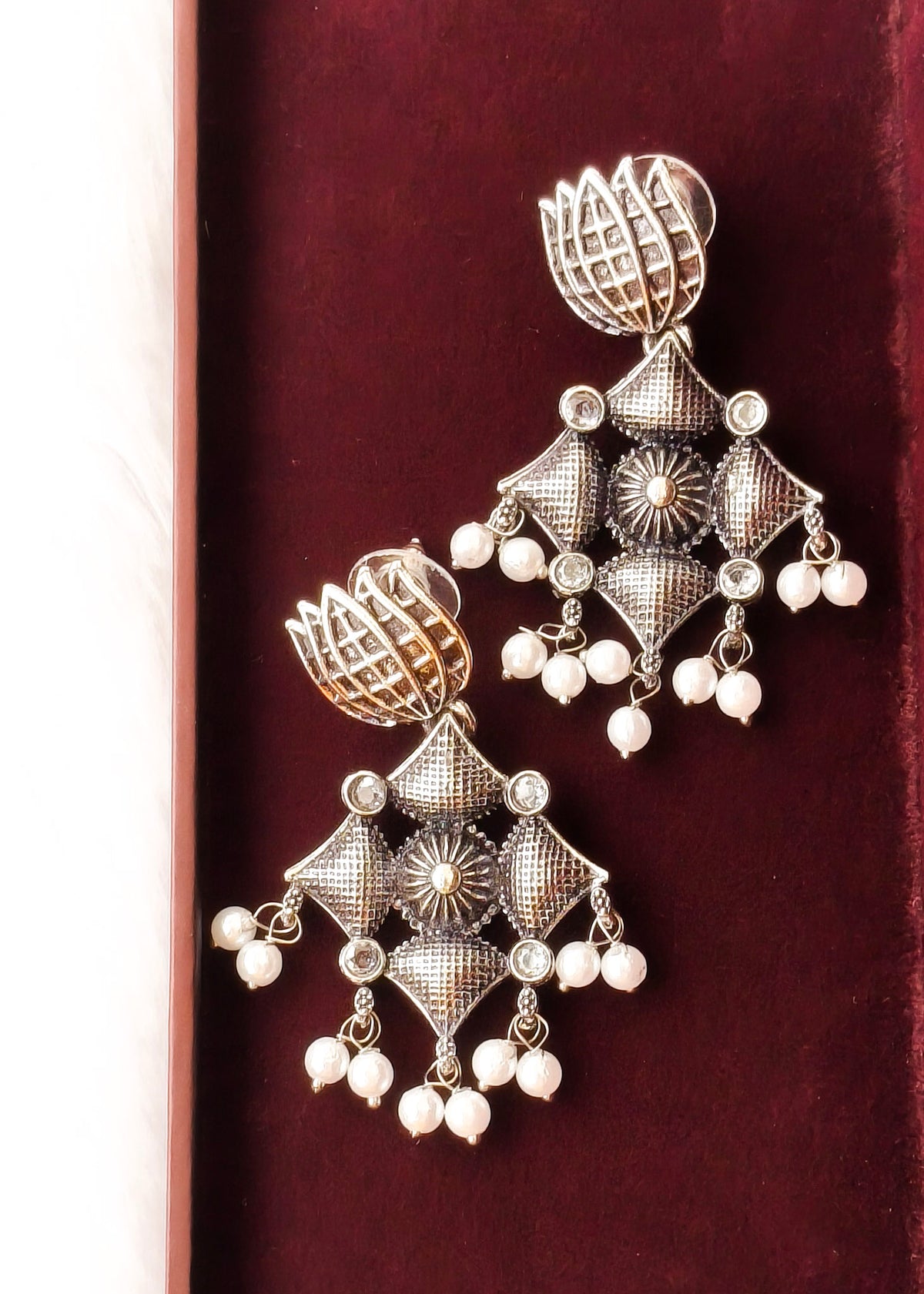 A pair of ornate silver-toned Indian jhumka earrings with intricate filigree patterns, small clear stones, and dangling white pearl beads, displayed on a dark red velvet surface.
