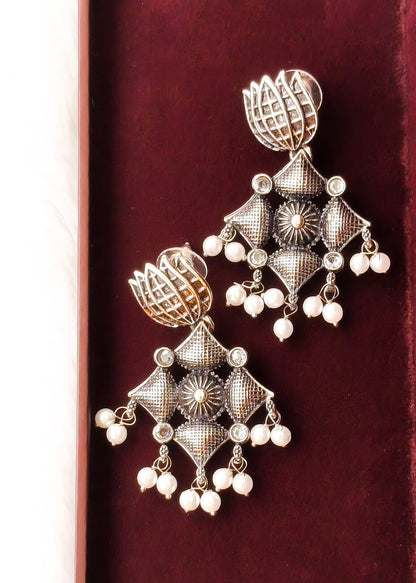 A pair of ornate silver-toned Indian jhumka earrings with intricate filigree patterns, small clear stones, and dangling white pearl beads, displayed on a dark red velvet surface.