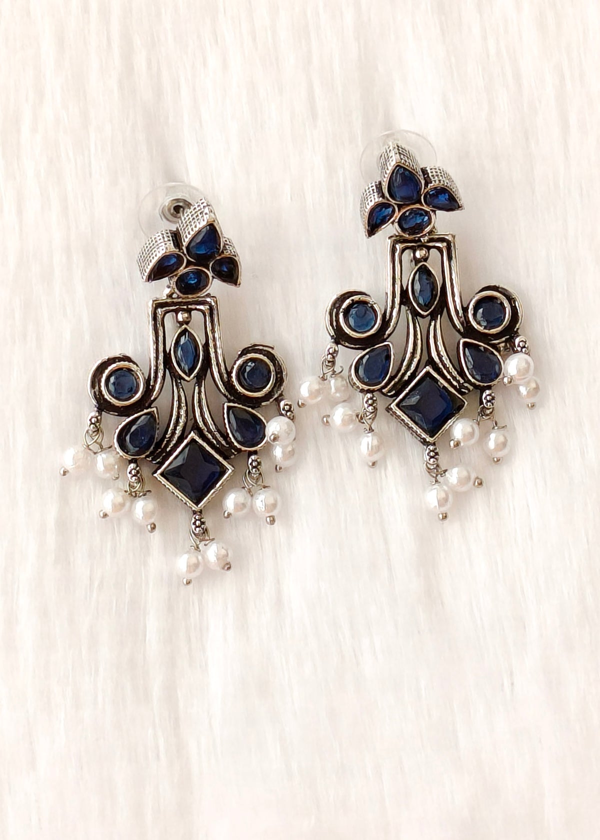 A pair of ornate silver-toned dangle earrings with dark blue stones and small white pearl drops, displayed on a soft white textured background.