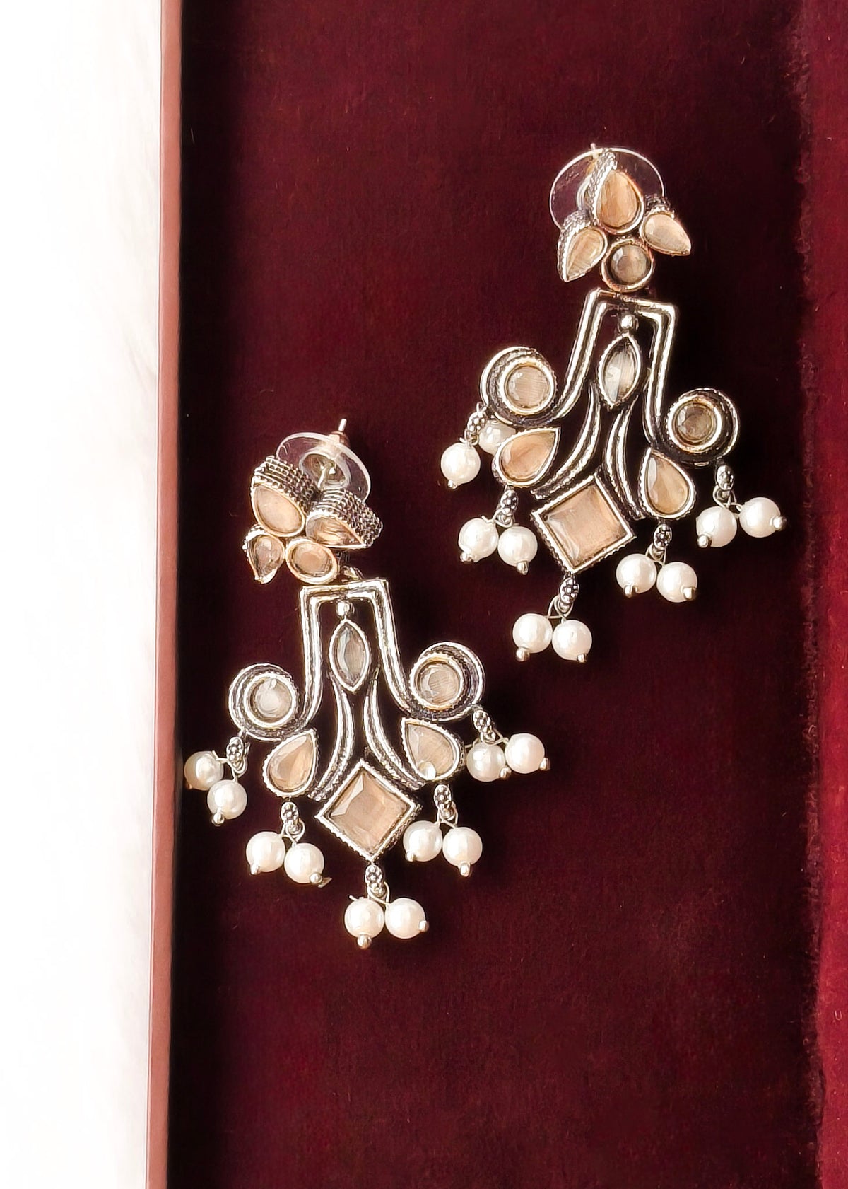 A pair of ornate silver-toned Indian chandelier earrings with cream-colored stones and white pearl drops, displayed inside a dark red velvet jewelry box.