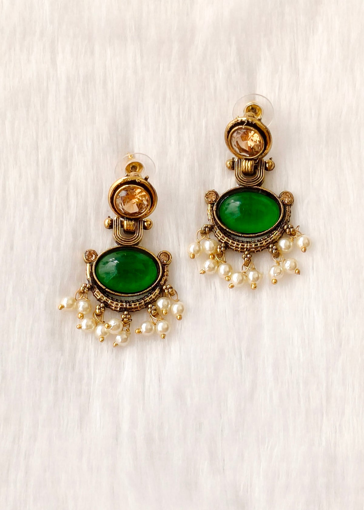 A pair of gold-toned statement earrings featuring oval green stones and dangling pearl-like beads, displayed on a light textured fabric background.