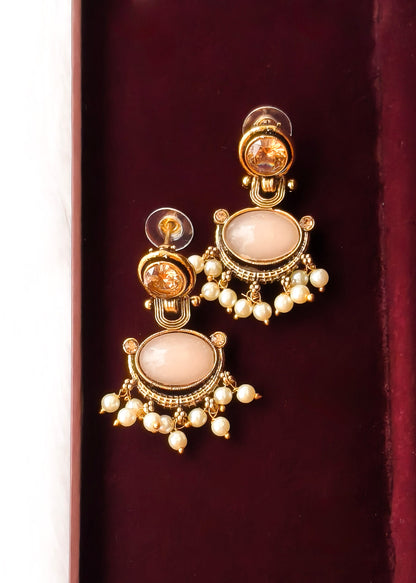 A pair of gold-plated statement earrings with large pale pinkish-beige oval stones and dangling pearl-like beads, displayed on a dark burgundy velvet surface.