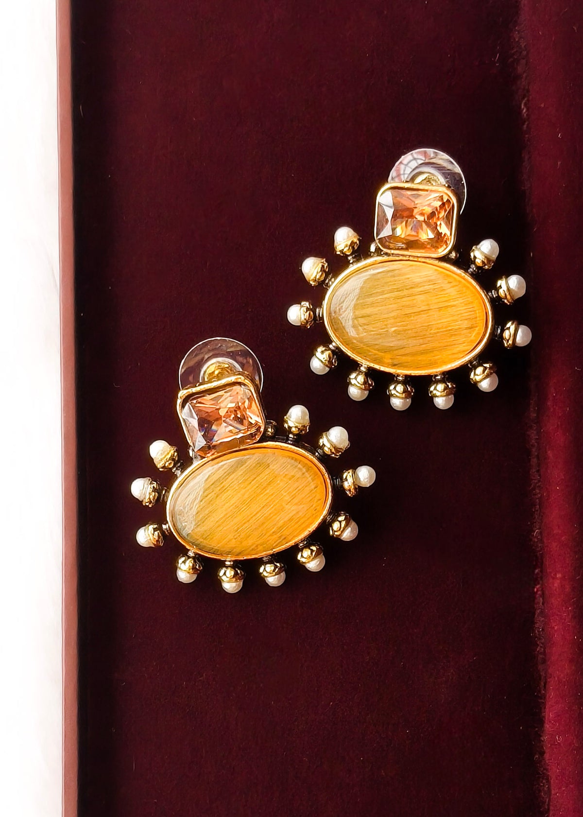 A pair of gold-toned statement earrings with oval amber-yellow centers, clear faceted top stones, and rows of small pearl-like beads around the edges, displayed on a dark burgundy velvet surface.