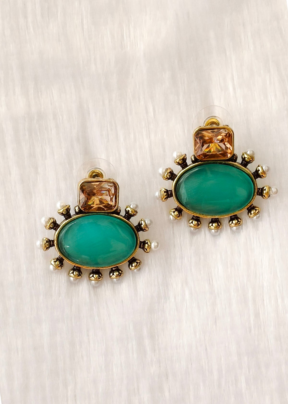 A pair of gold-toned statement earrings featuring large oval green cabochon stones and smaller square amber stones, accented with tiny white pearls, on a textured light gray background.