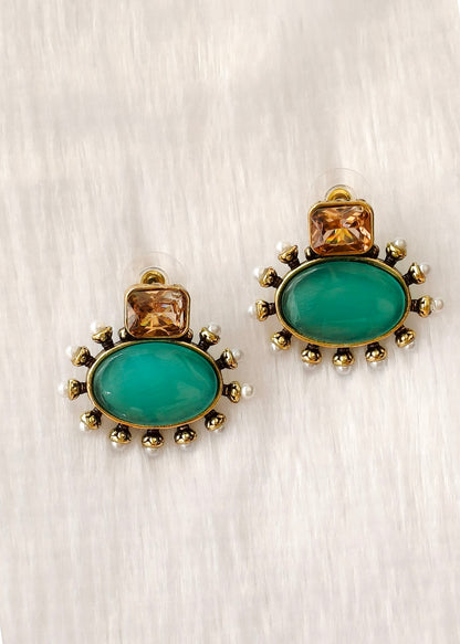 A pair of gold-toned statement earrings featuring large oval green cabochon stones and smaller square amber stones, accented with tiny white pearls, on a textured light gray background.