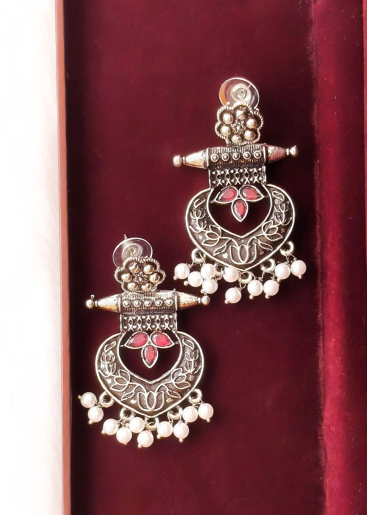 A pair of traditional Indian silver-toned jhumka earrings with red floral inlays and white bead drops, displayed on a deep red velvet surface.