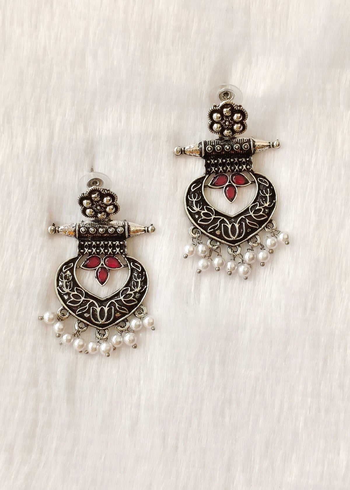 A pair of traditional Indian silver-toned dangle earrings with red floral accents and white pearl bead drops, displayed on a textured white fabric background.