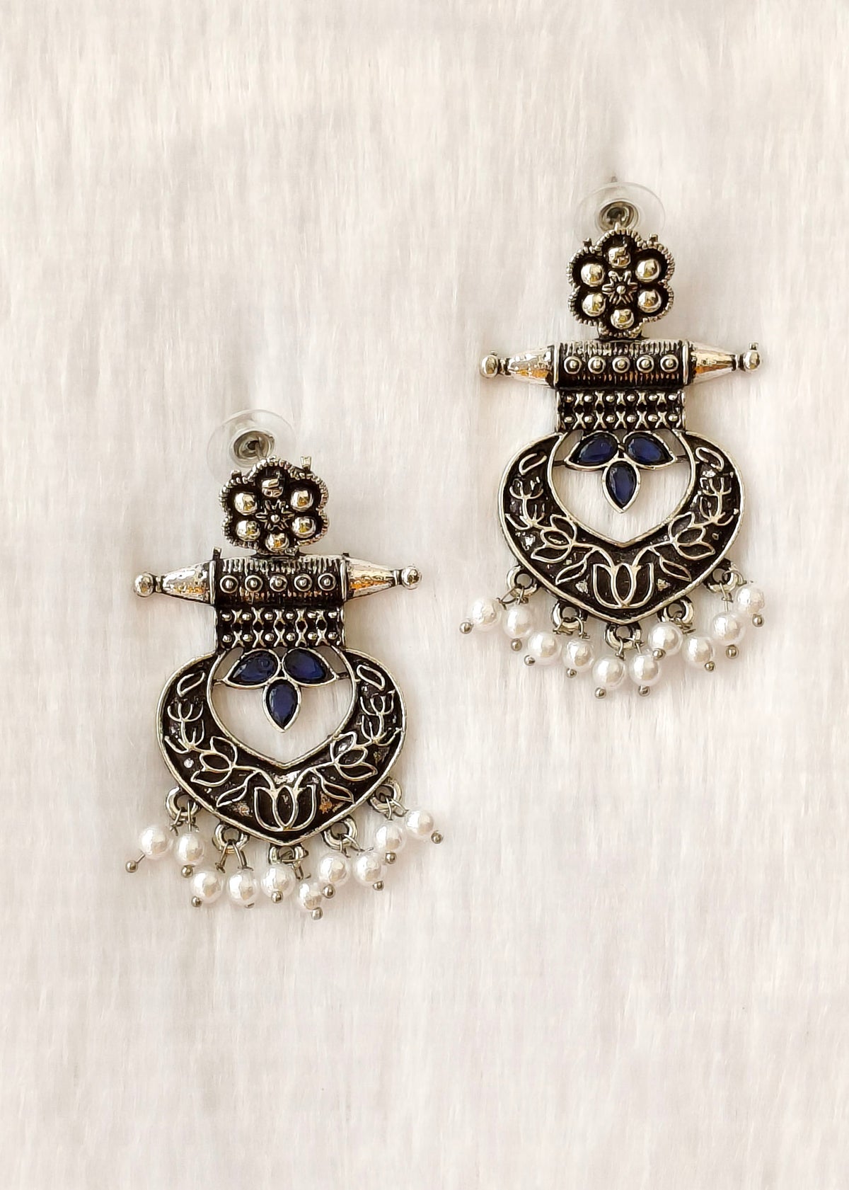 A pair of ornate silver-toned jhumka earrings with dark blue enamel details, floral motifs, and dangling pearl beads, displayed on a textured white fabric background.