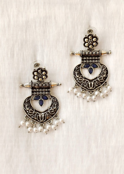 A pair of ornate silver-toned jhumka earrings with dark blue enamel details, floral motifs, and dangling pearl beads, displayed on a textured white fabric background.