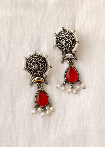 A pair of traditional Indian silver-toned dangle earrings with red teardrop stones and small white pearl bead drops, displayed on a textured off-white fabric background.