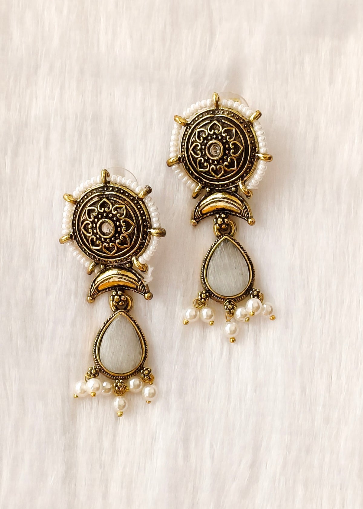 A pair of ornate drop earrings with antique gold-toned metalwork, white faux pearls, and teardrop-shaped white stones, displayed on a soft white textured fabric background.