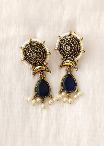A pair of ornate gold-toned dangle earrings with circular filigree tops, white pearl bead borders, and dark navy blue teardrop-shaped stones, displayed on a soft white textured fabric background.