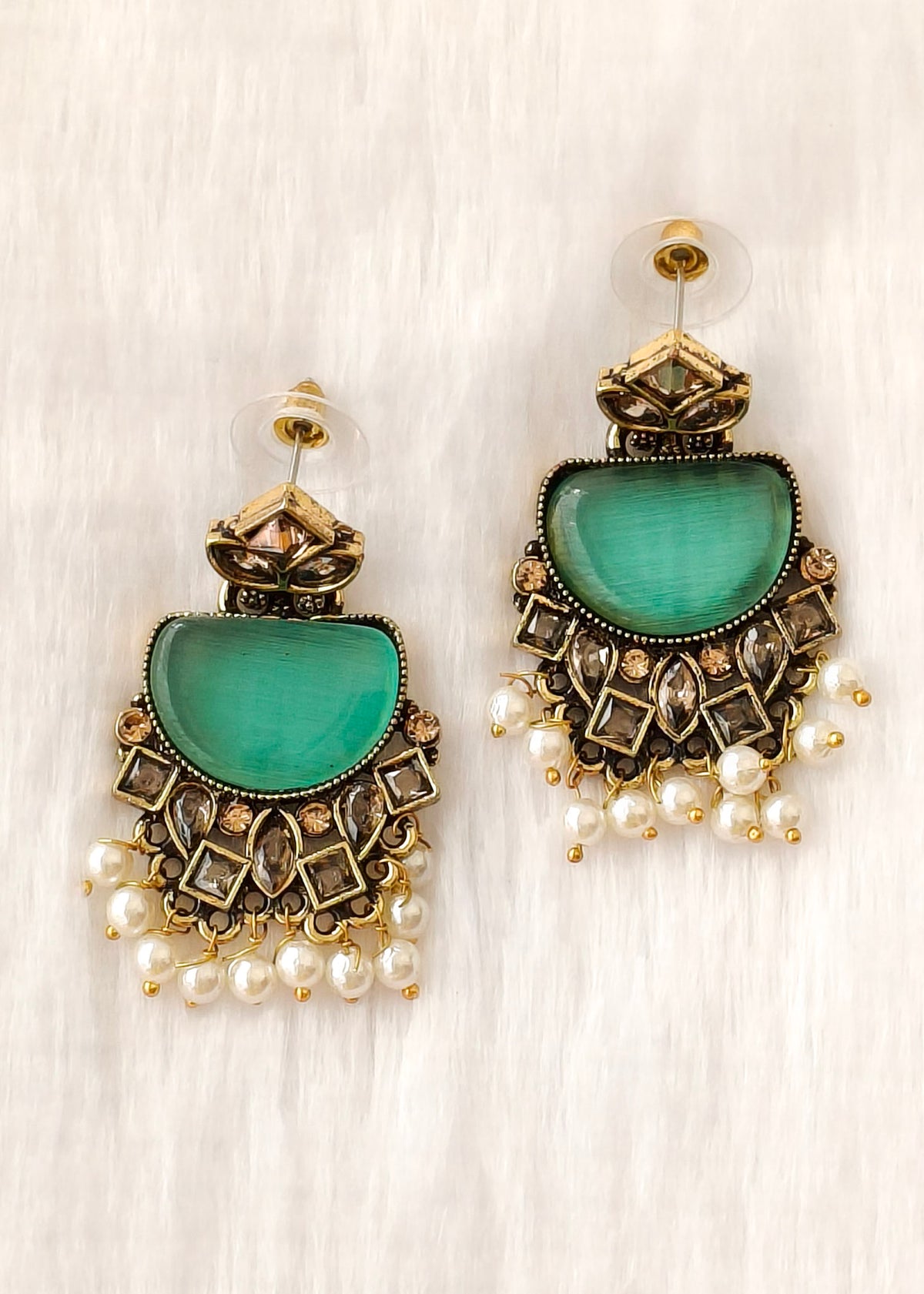 A pair of ornate gold-toned statement earrings featuring a large green semicircle stone, intricate geometric detailing, and dangling pearl beads, displayed on a white textured background.