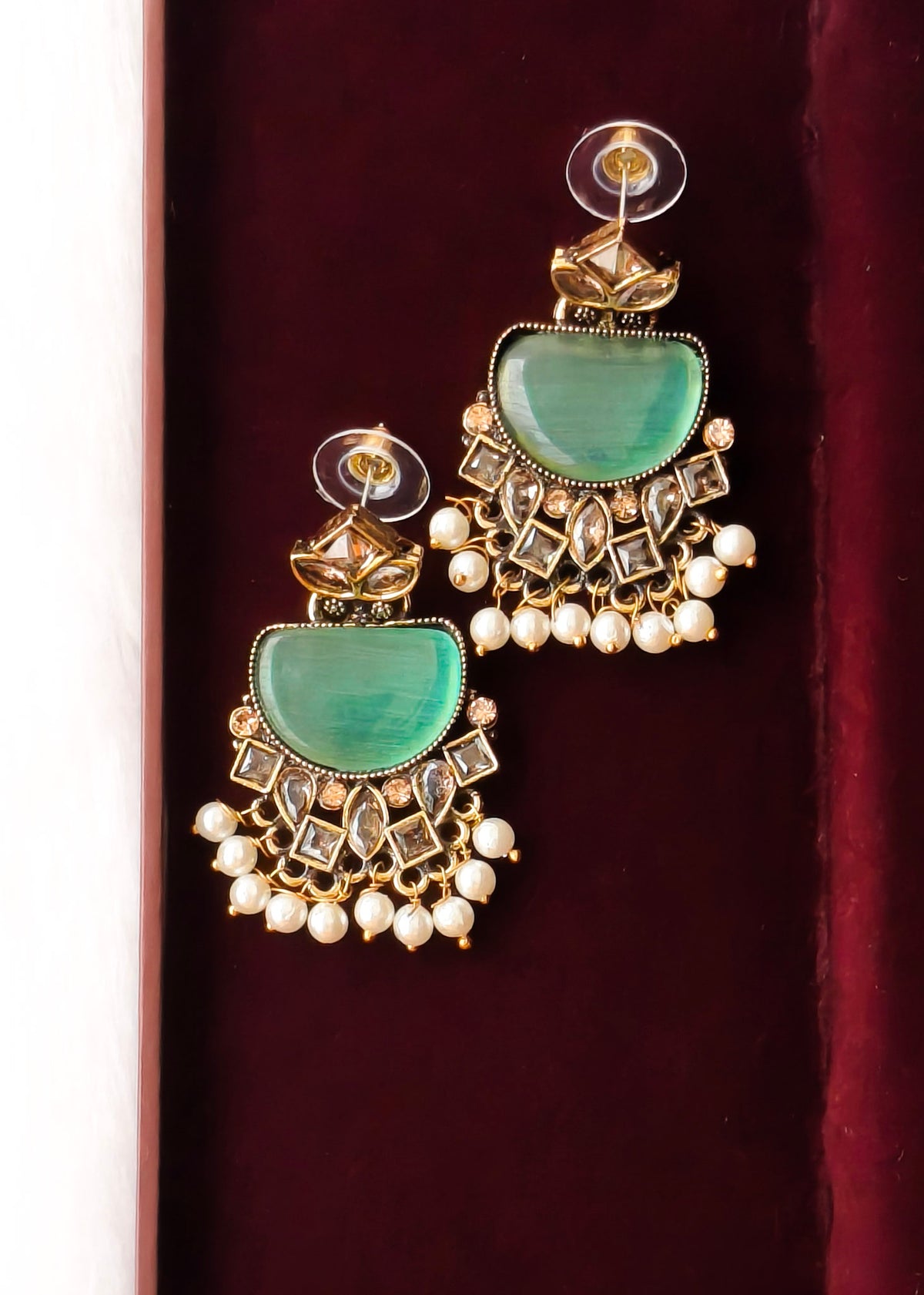 A pair of gold-toned traditional Indian jhumka earrings with green stone inlays and white pearl bead fringe, displayed on a dark red velvet surface.