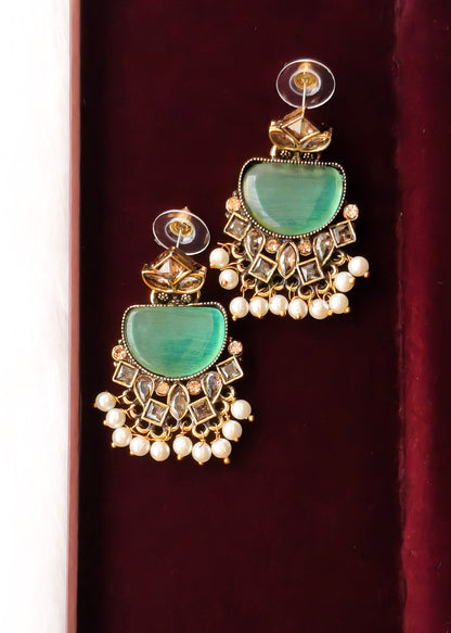 A pair of gold-toned traditional Indian jhumka earrings with green stone inlays and white pearl bead fringe, displayed on a dark red velvet surface.