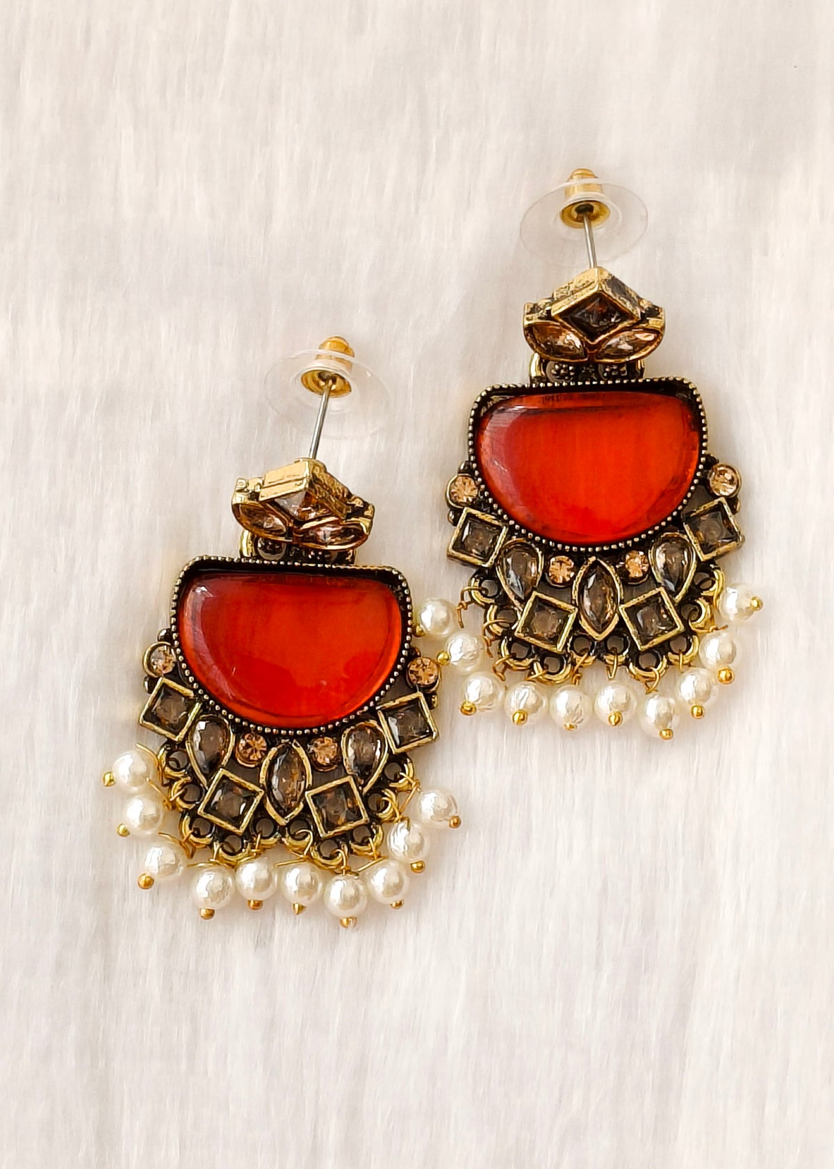 A pair of ornate gold-toned statement earrings with large oval red-orange cabochon stones, intricate geometric metalwork, and dangling pearl beads, displayed on a white textured fabric background.