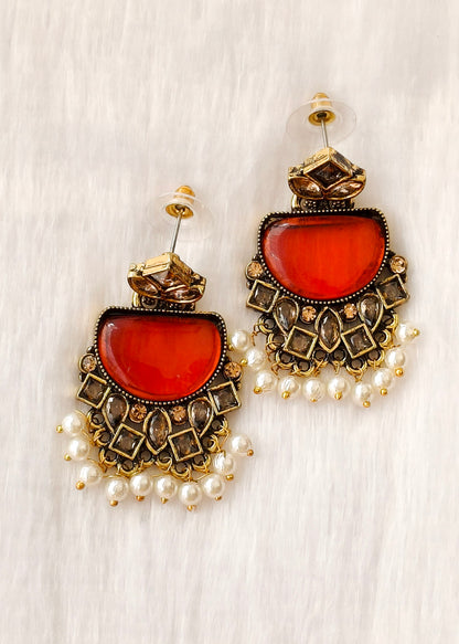 A pair of ornate gold-toned statement earrings with large oval red-orange cabochon stones, intricate geometric metalwork, and dangling pearl beads, displayed on a white textured fabric background.