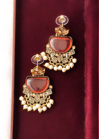 A pair of ornate gold-toned Indian jhumka earrings with large oval amber-red stones, clear rhinestone accents, and dangling pearl-like beads, displayed on a deep burgundy velvet surface.