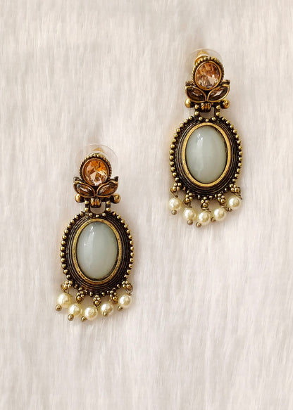 A pair of antique bronze drop earrings with oval light green stone centers, small amber crystal accents at the top, and dangling pearl beads at the bottom, displayed on a soft white textured fabric background.