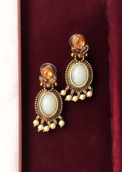 A pair of gold-plated traditional Indian dangle earrings with oval white stone centers and small pearl-like bead drops, displayed on a dark red velvet surface.