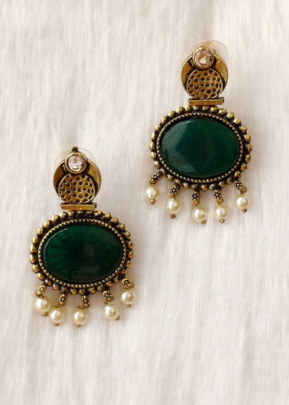 A pair of gold-plated statement earrings featuring oval dark green stones, beaded borders, and dangling pearl-like beads, displayed on a textured white fabric background.