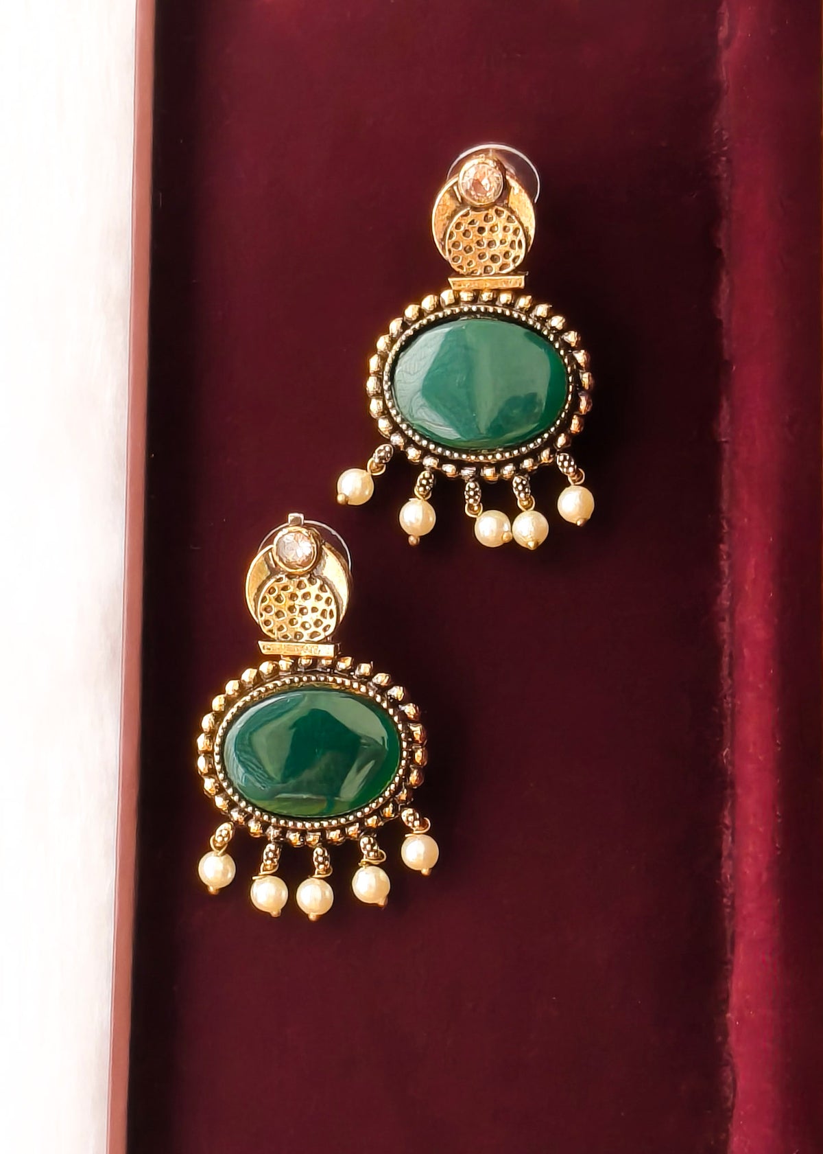 A pair of gold-plated traditional Indian earrings with oval green stone centers and dangling pearl-like beads, displayed on a dark red velvet surface.