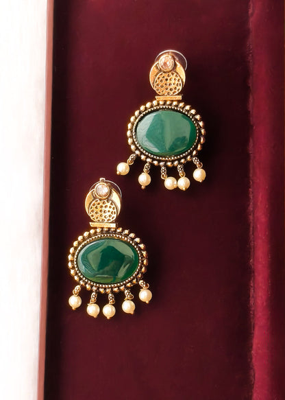 A pair of gold-plated traditional Indian earrings with oval green stone centers and dangling pearl-like beads, displayed on a dark red velvet surface.