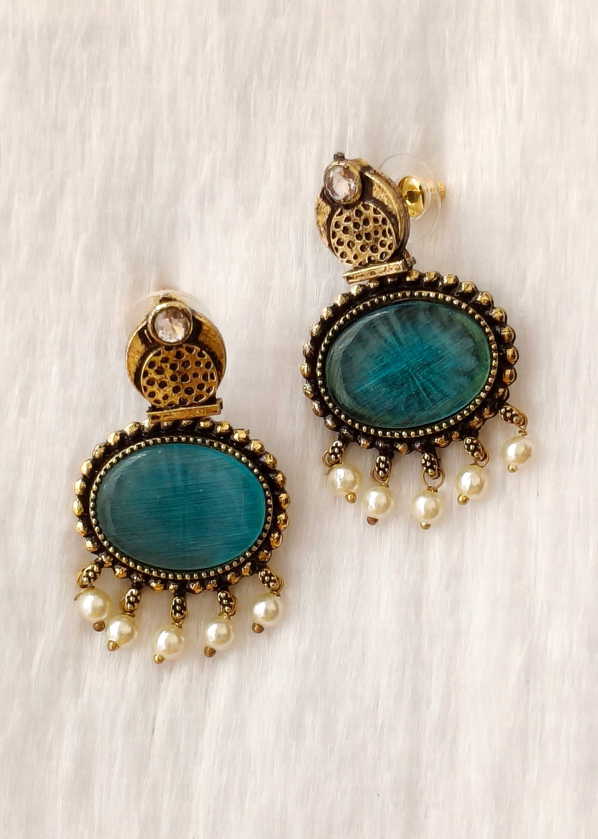 A pair of ornate drop earrings with oval turquoise-blue stones, antique gold-tone filigree tops, and dangling pearl beads, displayed on a soft white textured fabric background.