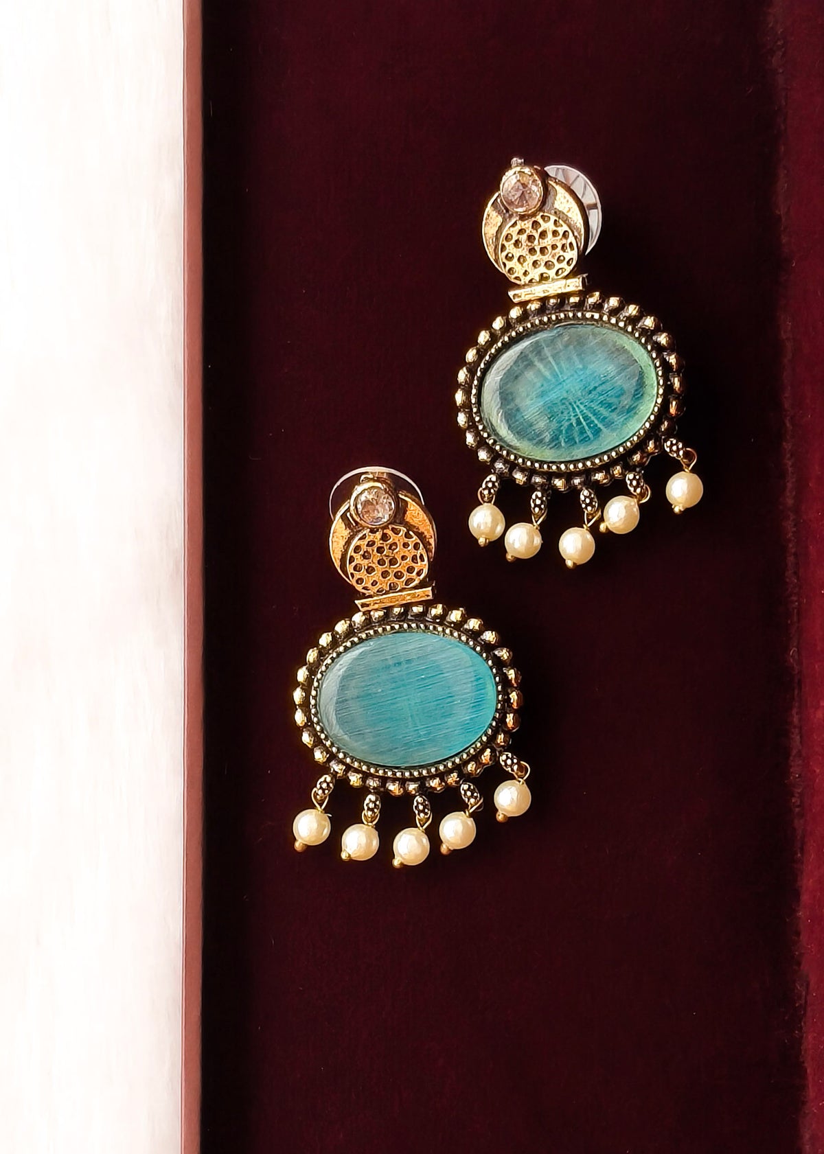 A pair of ornate gold-toned dangle earrings with oval turquoise-blue stones and small pearl-like drops, displayed on a dark burgundy velvet surface.