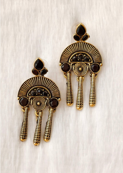 A pair of gold-toned statement earrings with dark brown enamel details and small black stones, displayed on a textured white background.