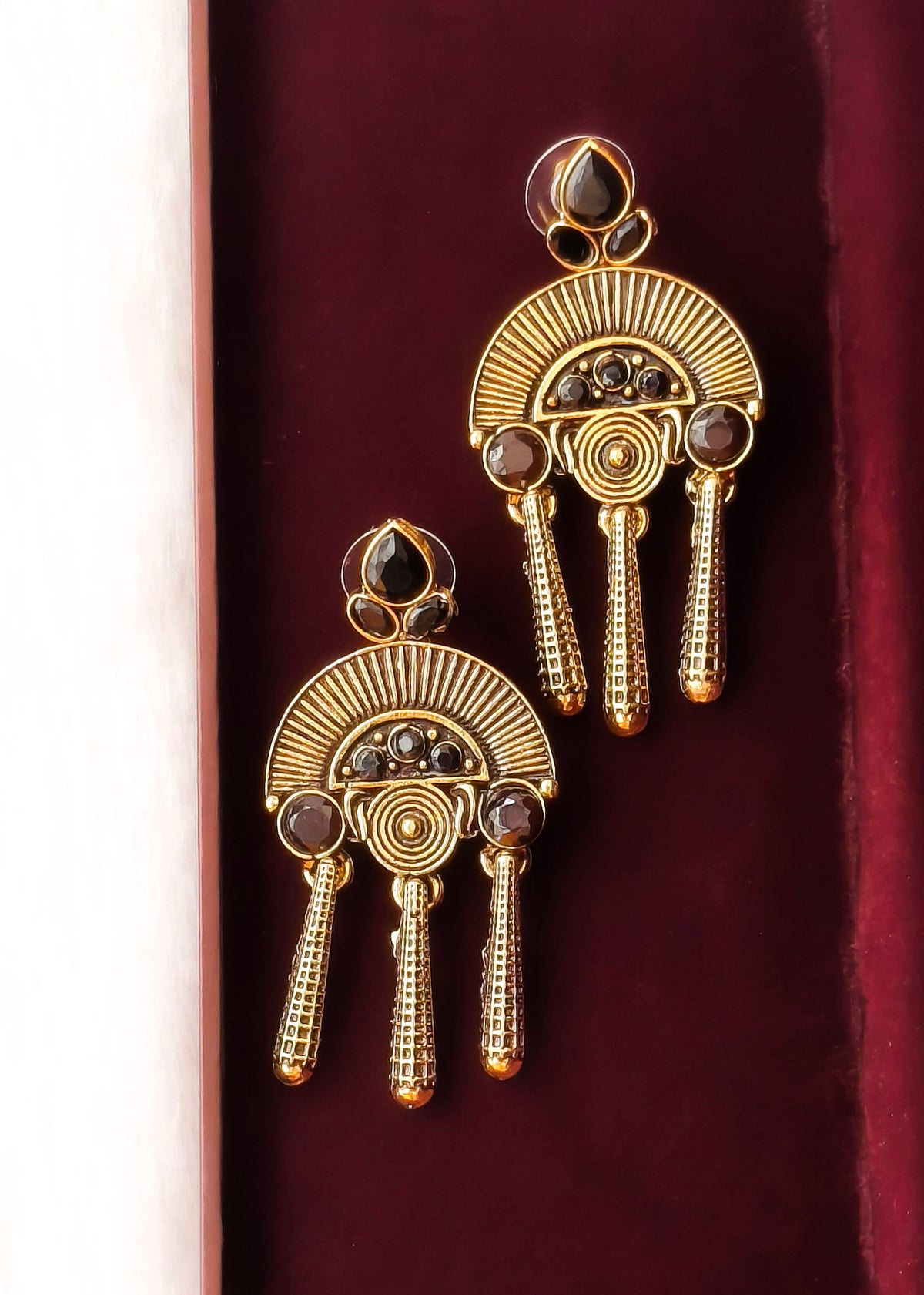 A pair of ornate gold-toned statement earrings with fan-shaped sunburst detailing, circular spiral motifs, and dangling textured chains, displayed on a deep burgundy velvet surface.