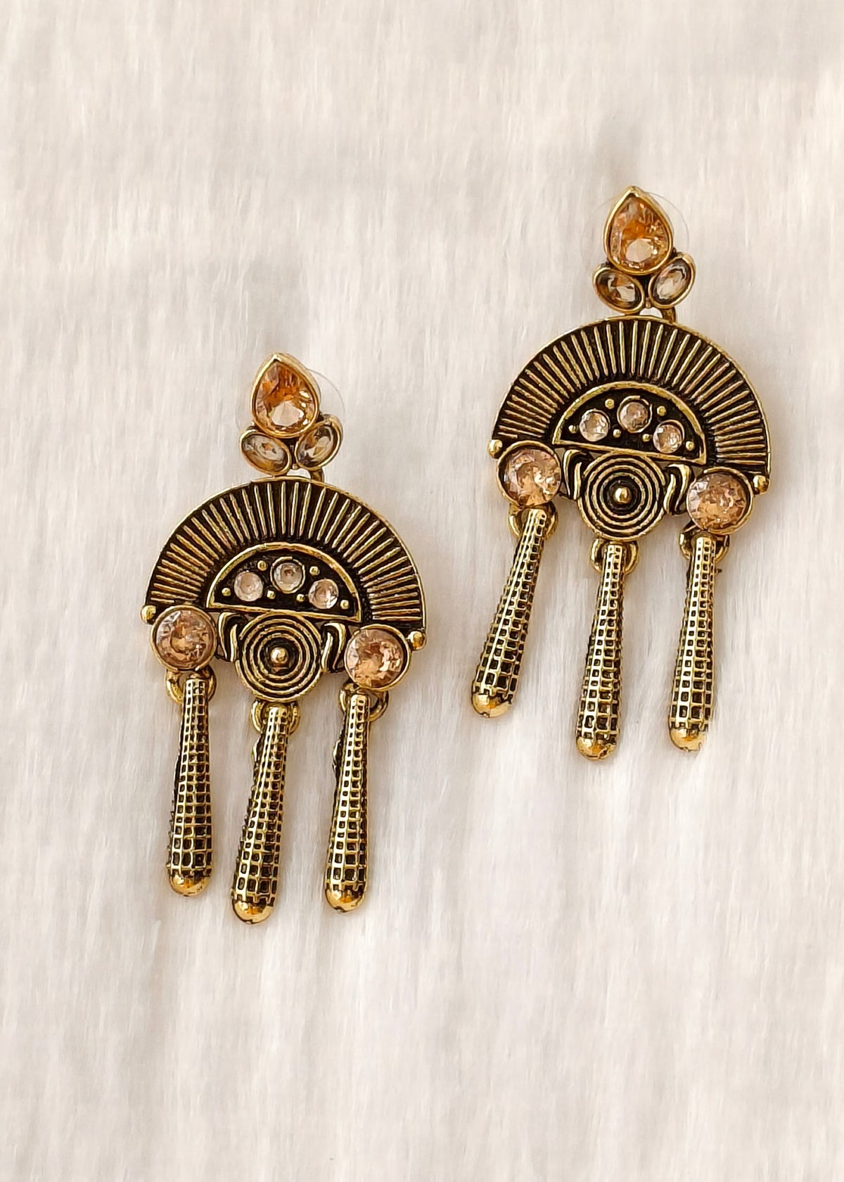 A pair of gold-toned statement earrings with a fan-shaped sunburst design, small clear stones, and three dangling textured chains, displayed on a textured off-white background.