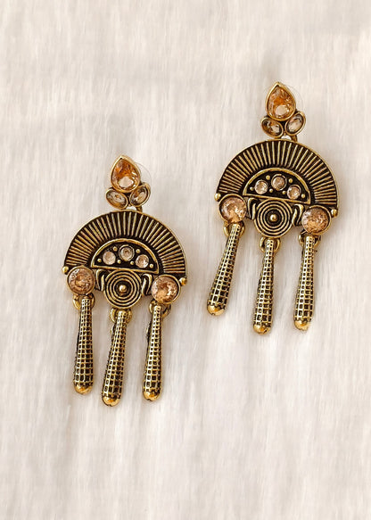 A pair of gold-toned statement earrings with a fan-shaped sunburst design, small clear stones, and three dangling textured chains, displayed on a textured off-white background.