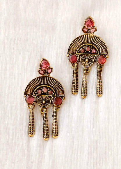 A pair of ornate gold-toned statement earrings with pink gemstone accents, displayed on a textured white background.