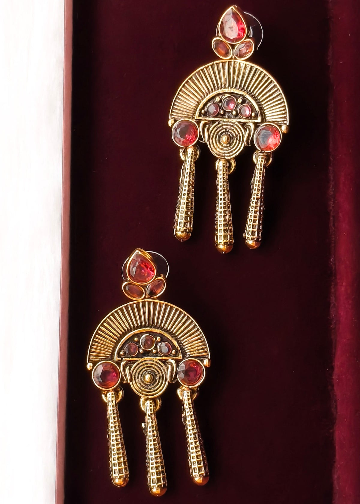 A pair of gold-toned traditional Indian earrings with red and pink stones, displayed on a dark red velvet surface.