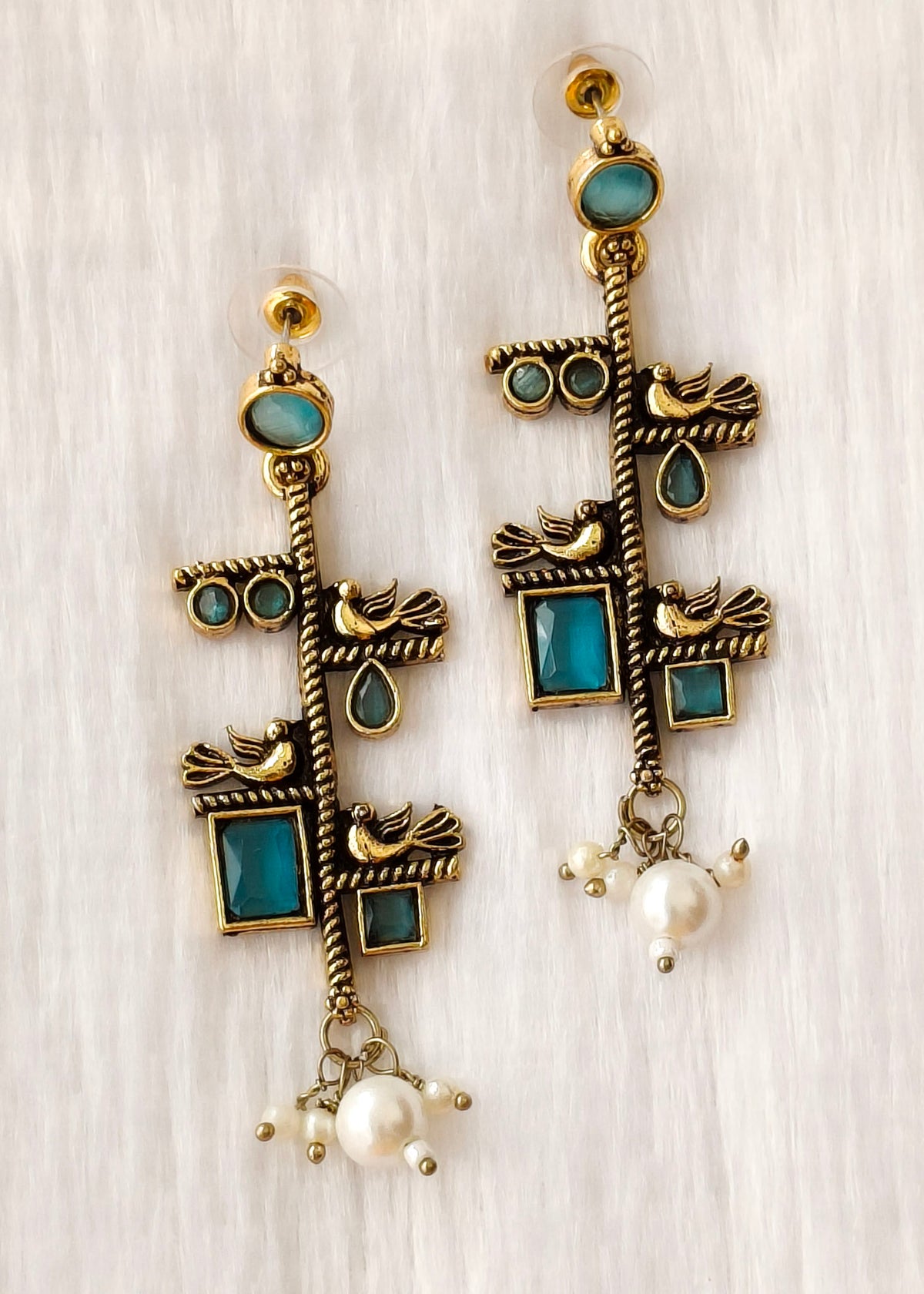 A pair of bohemian-style gold-plated dangle earrings with turquoise blue stone inlays, small bird motifs, and dangling white pearl beads, displayed on a light wood background.
